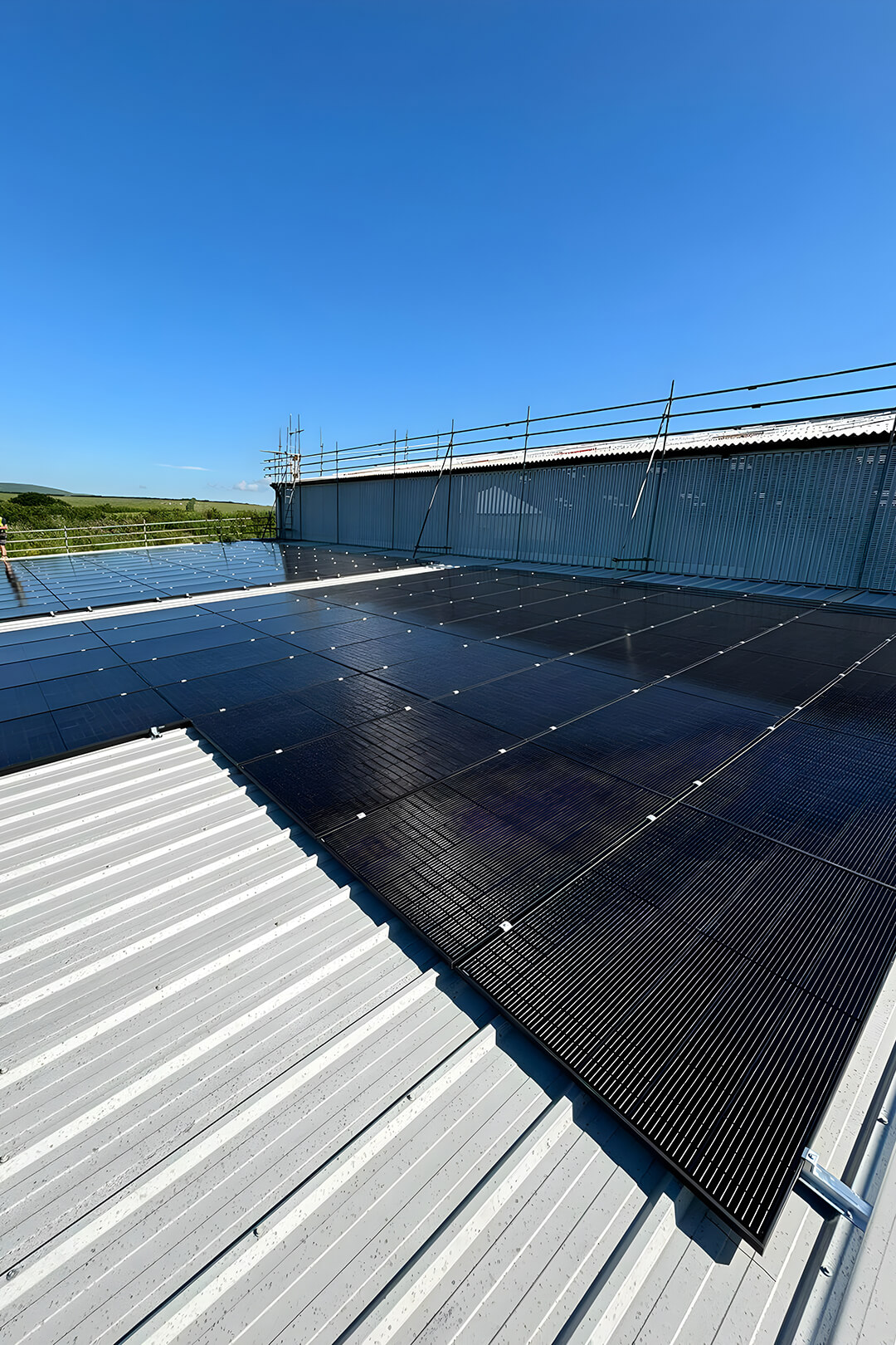 Rooftop solar panels under clear blue sky on a large metal building. Panels are neatly aligned, with distant green hills and scaffolding visible.